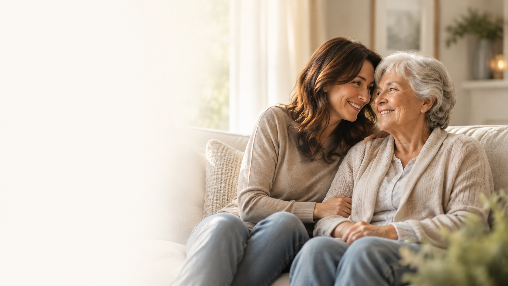 A woman leaning her head against her elderly mother as they sit together on a sofa in a sunlit living room